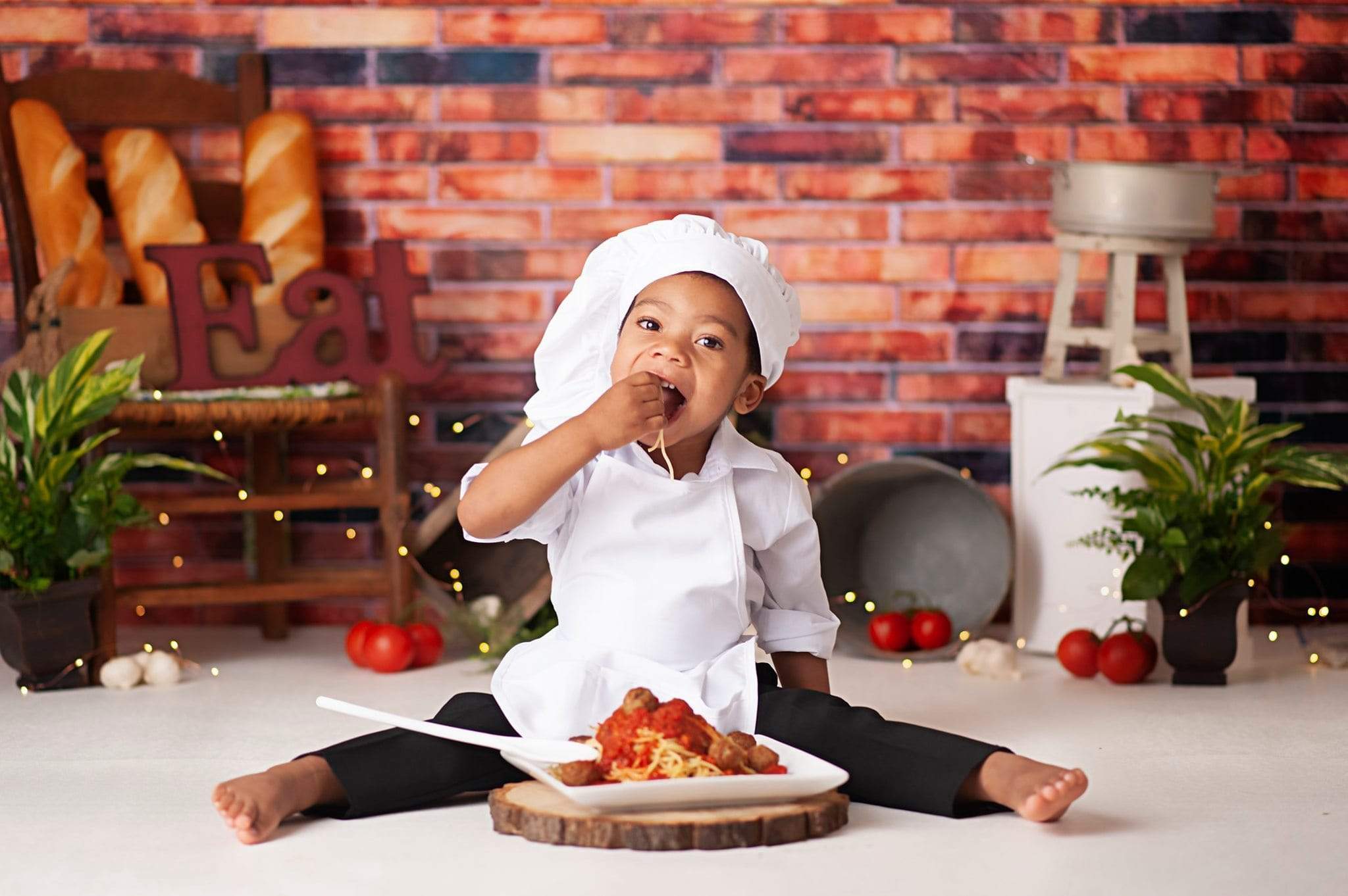 Boy in chef costume eating noodles in front of red brick wall backdrop
