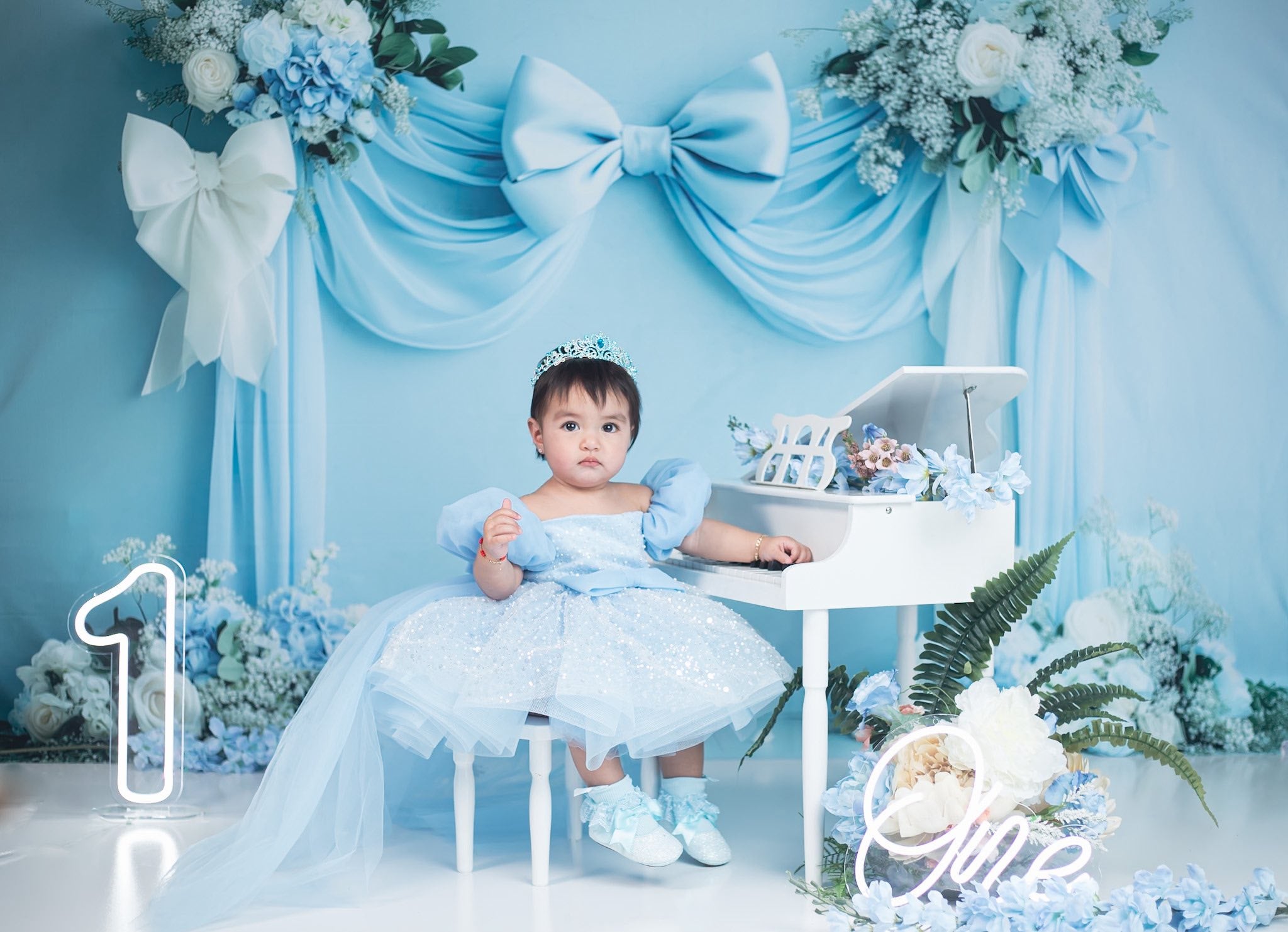 A little girl sits in front of a blue bow backdrop, playing the piano.