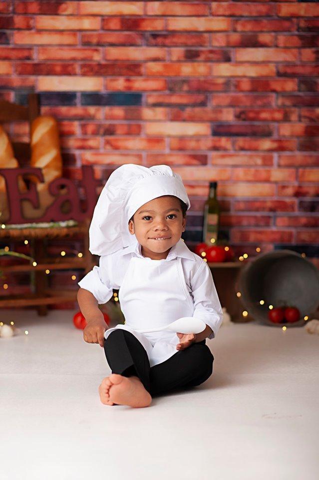 Boy in chef costume eating noodles in front of red brick wall backdrop